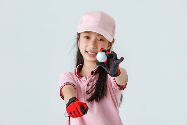 閉じるアップ隔離されたスタジオショットのゴルフボールの手のアジアの小さなプロの女の子の子供のゴルファーでピンクスポーツファッション服赤手袋とキャップスタンドパターを目指している緑の草. - 写真・画像