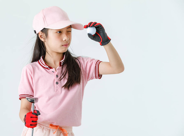 Close up isolated studio shot of Asian little professional girl kid golfer in pink athlete fashion clothing red gloves and cap stand hold driver and golf ball close eye aiming for target on green. - Photo, Image