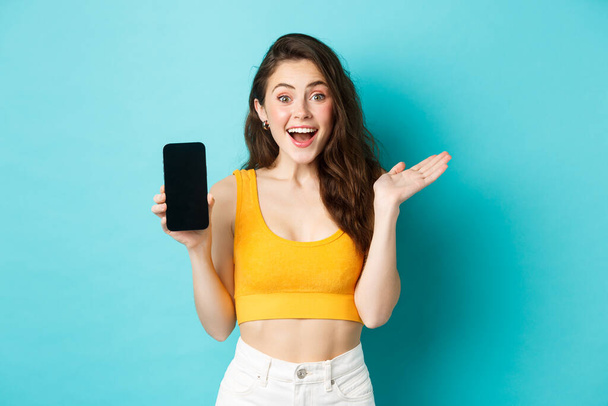 Attraktive junge Frau in Glamour-Sommerkleidung, zeigt leeren Smatphone-Bildschirm, keucht erstaunt, empfiehlt App, steht vor blauem Hintergrund - Foto, Bild
