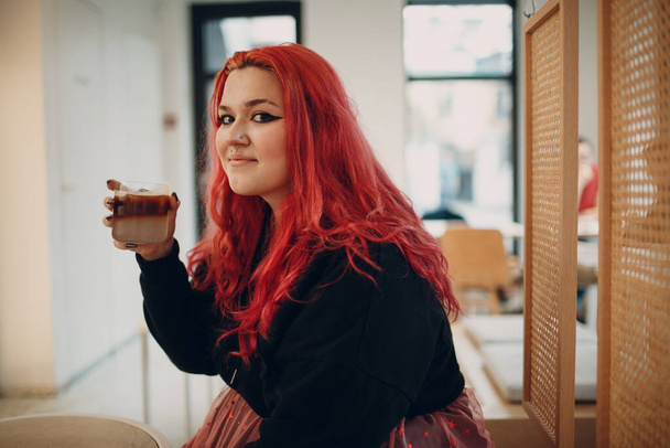 Donna europea plus size. Giovane ragazza rossa dai capelli rosa corpo positivo seduto in caffè con bevanda di caffè. - Foto, immagini
