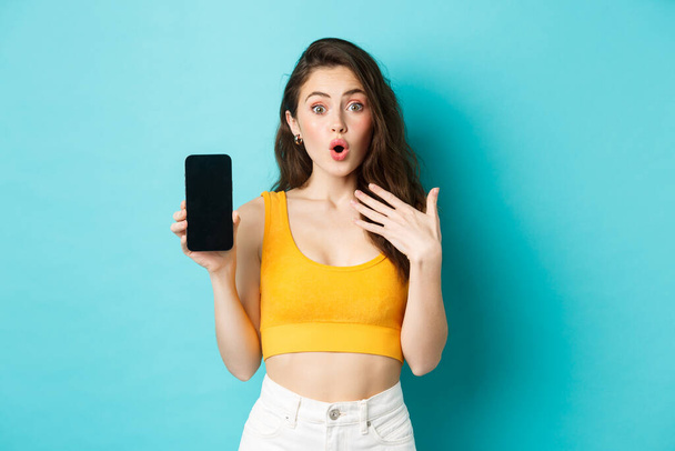 Porträt eines überraschten Mädchens beeindruckt mit Smartphone-App, zeigt leeren Handy-Bildschirm und sieht erstaunt aus, steht über blauem Hintergrund - Foto, Bild