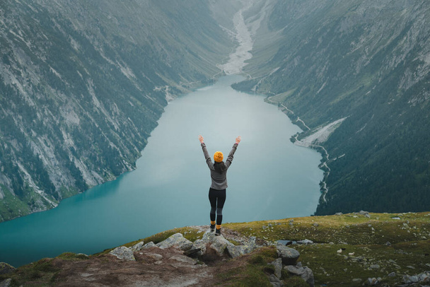 Женщина-туристка празднует успех на вершине горы с распростертыми объятиями и великолепным видом над бирюзовым горным озером. Радостно приветствуя успешную молодую женщину-альпинистку с руками, устремленными к вершине - Фото, изображение