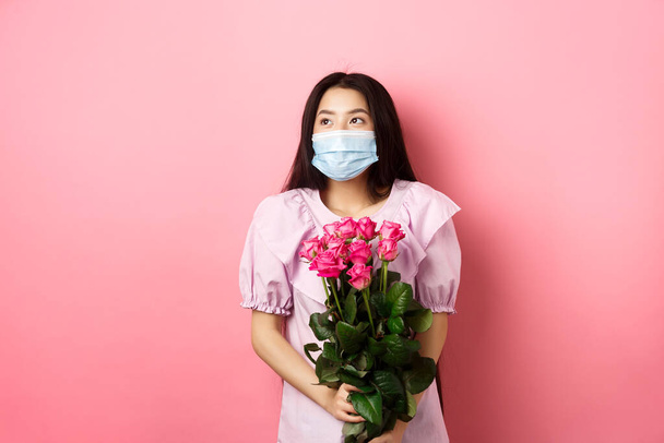 Romantyczna Azjatka w masce medycznej spoglądająca na pustą przestrzeń z marzycielskimi oczami, trzymająca bukiet róż na Walentynki, umawiająca się z kochankiem podczas pandemii - Zdjęcie, obraz