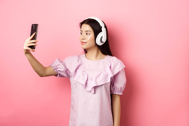 Schöne Japanerin macht Selfie auf Smartphone, trägt drahtlose Kopfhörer, steht vor rosa Hintergrund - Foto, Bild