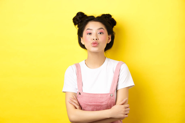 Cute teen asian girl pucker lips for kiss, cross arms on chest and look romantic at camera, yellow background - Photo, Image