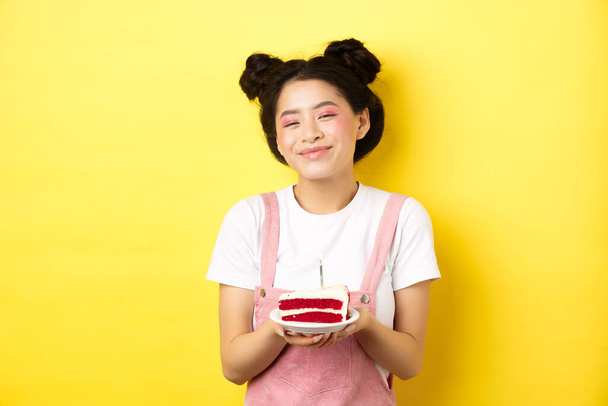 feliz asiático cumpleaños chica con brillante maquillaje, soplando vela en pastel, haciendo deseo, de pie sobre amarillo fondo - Foto, Imagen