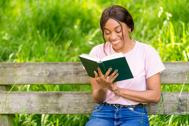 Giovane donna nera seduta su una panchina del parco a leggere un libro. - Foto, immagini