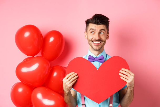Valentines day concept. Romantic guy smiling and looking left, dreaming of date with lover, showing red big heart cutout, pink background - Foto, imagen