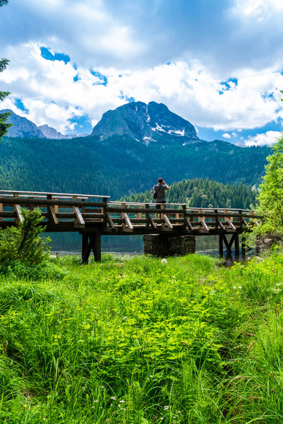 Tourist man on wooden footbridge over stream. Meded Peak and Glacial Black Lake. Durmitor National Park. Walking path near lake is a popular destination for recreation and hiking. Montenegro. - Photo, Image
