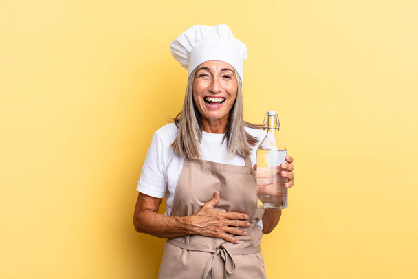 middle age chef woman laughing out loud at some hilarious joke, feeling happy and cheerful, having fun with a water bottle - Foto, Imagen