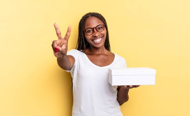smiling and looking happy, carefree and positive, gesturing victory or peace with one hand and holding an empty box - Photo, image
