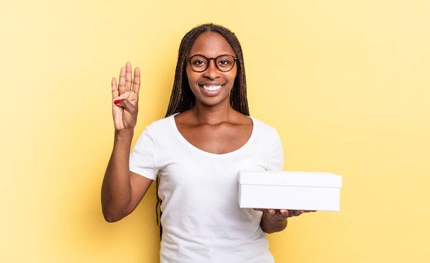 smiling and looking friendly, showing number four or fourth with hand forward, counting down and holding an empty box - Foto, afbeelding