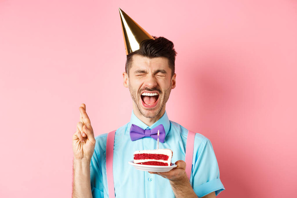 Ünnepek és ünnepség koncepció. Izgalmas srác ünnepli születésnapját és kívánság, keresztujjú szerencsét, miközben tartja bday torta gyertya, rózsaszín háttér - Fotó, kép
