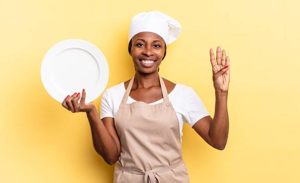 black afro chef woman smiling and looking friendly, showing number four or fourth with hand forward, counting down. empty plate concept - Φωτογραφία, εικόνα