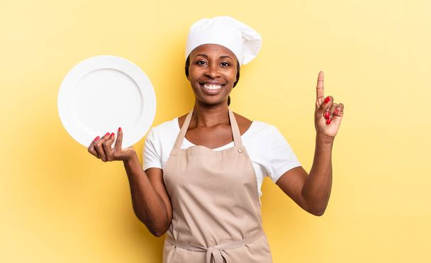 black afro chef woman smiling and looking friendly, showing number one or first with hand forward, counting down. empty plate concept - Φωτογραφία, εικόνα