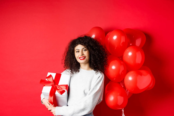 Beautiful caucasian woman with dark curly hair and red lips, standing with romantic hearts balloons and present in box, receive gift from lover, standing on studio background - Photo, Image