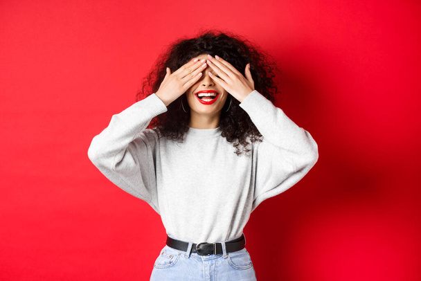 Happy beautiful woman with curly hair and red lips, covering eyes with hands and waiting surprise, smiling excited, standing against red background - Foto, Imagen