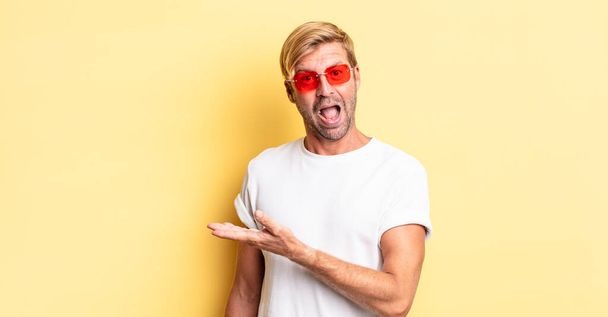 blond adult man smiling cheerfully, feeling happy and showing a concept and wearing sunglasses - Foto, afbeelding