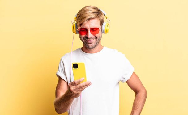 blond adult man smiling happily with a hand on hip and confident with headphones - Fotó, kép
