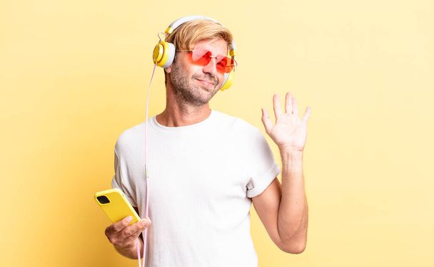 blond adult man smiling happily, waving hand, welcoming and greeting you with headphones - Foto, imagen
