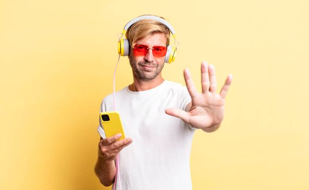 blond adult man smiling and looking friendly, showing number five with headphones - Foto, immagini