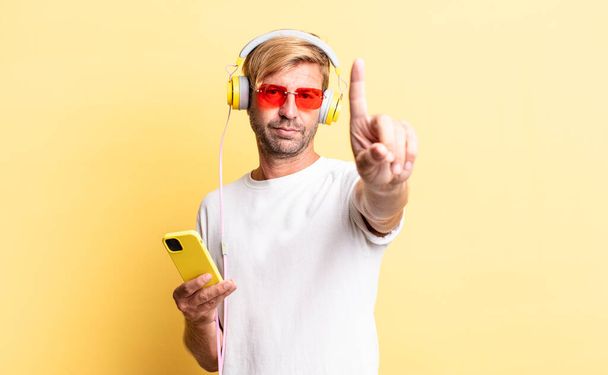 blond adult man smiling and looking friendly, showing number one with headphones - Φωτογραφία, εικόνα