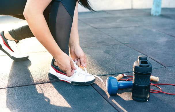 a young girl bent down to tie her shoelaces against the background of a dumbbell a jump rope and a water bottle. - Photo, Image