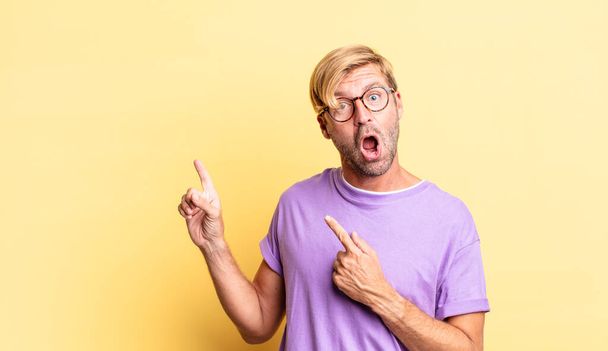 handsome blond adult man feeling joyful and surprised, smiling with a shocked expression and pointing to the side - Foto, imagen