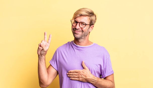 handsome blond adult man looking happy, confident and trustworthy, smiling and showing victory sign, with a positive attitude - Foto, afbeelding