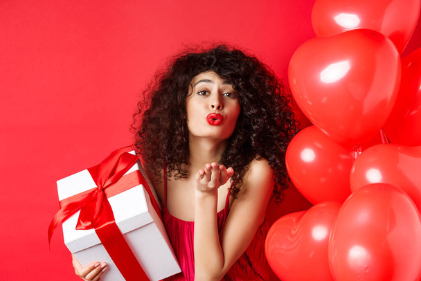 Romance and Valentines day concept. Pretty curly-haired girl in red dress sending her love, blowing air kiss at camera, holding gift from lover, standing near hearts on red background - Photo, Image