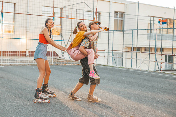 Group of friends hanging out outside. One girl back riding her friend, other rides behind on rollers. Positive vibes - Foto, imagen