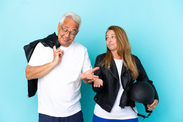 Mavi arka planda izole edilmiş motosiklet kasklı orta yaşlı çiftler omuzlarını kaldırırken önemsiz jestler yapıyorlar. - Fotoğraf, Görsel