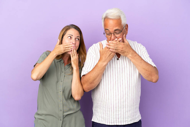 Пара средних лет изолирована на фиолетовом фоне, прикрывая рот руками - Фото, изображение