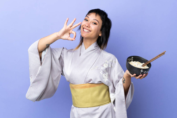 Mujer joven que usa kimono sobre un fondo azul aislado mostrando un signo aceptable con los dedos mientras sostiene un tazón de fideos con palillos - Foto, Imagen