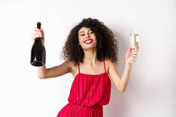 赤いドレスの幸せなパーティーの女の子、シャンパンとガラスのボトルで踊る、飲みながら楽しむ、休日を祝う、白い背景に立って - 写真・画像
