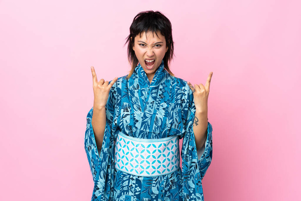 Jonge vrouw draagt kimono over geïsoleerde blauwe achtergrond maken hoorn gebaar - Foto, afbeelding