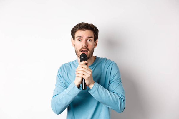 verwarde jongeman die zenuwachtig naar de camera kijkt terwijl hij karaoke zingt, microfoon vasthoudt, over een witte achtergrond staat - Foto, afbeelding