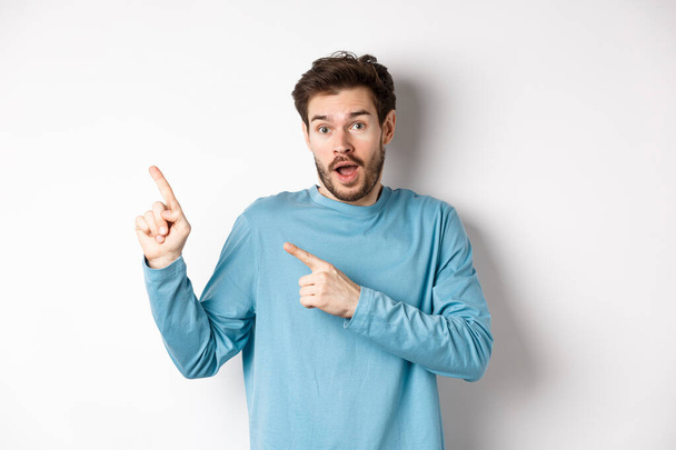 Amazed man showing awesome promo offer, pointing fingers at upper left corner logo and looking excited, recommend product, white background - Photo, Image