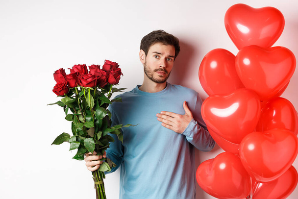 Romanttinen kaveri ilmaista rakkautensa Ystävänpäivä lahjoja, tuo kimppu punaisia ruusuja ja ilmapalloja, pitämällä käsi sydämellä, seisoo valkoisella taustalla - Valokuva, kuva