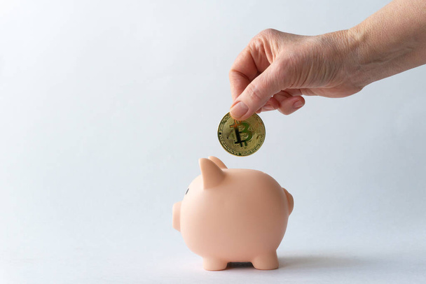 Mão de mulher caucasiana idosa cai moeda bitcoin metal em porquinho banco rosa porco no fundo branco com lugar para colocar texto. Finanças domésticas - Foto, Imagem