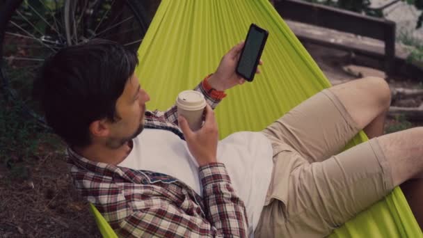 Man browses Internet on smartphone while drinking coffee with him while sitting in green hammock overlooking lake in a forest park having arrived by bike. Cyclist in hammock at campsite by river - Footage, Video