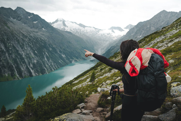 Backpacking and trekking tourism concept. Side view of female hiker tourist pointing hand on right direction. Young woman with backpack and trekking poles looking for directions in Zillertal, Austria - Photo, Image