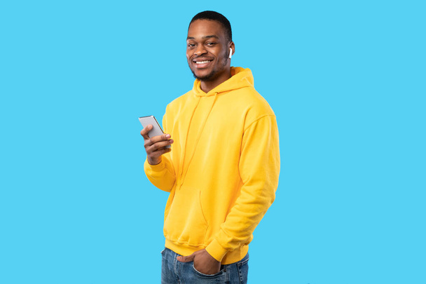 African American Guy Using Smartphone Wearing Earbuds Over Blue Background - Fotó, kép