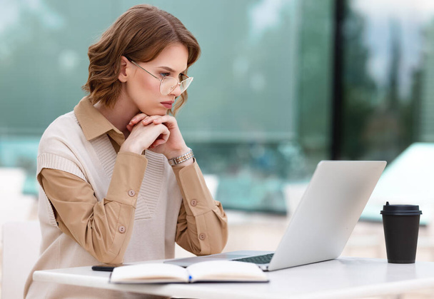 Серйозна концентрована молода жінка в окулярах працює дистанційно на ноутбуці онлайн, сидячи з кавою на терасі на відкритому повітрі в кафе, роблячи нотатки час від часу у відкритому порядку
 - Фото, зображення