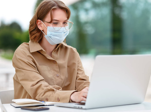 Young female entrepreneur in medical mask holding video conference call with partner or employees, tutor giving online lesson sitting outside with laptop. Remote work during quarantine concept - Foto, imagen