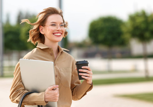 Nahaufnahme Foto von entzückenden jungen Mädchen Bürosekretärin zu Fuß nach draußen mit strahlendem breitem Lächeln, während sie Tasse Kaffee zum Mitnehmen und Laptop vor dem Hintergrund der städtischen Außen in sonnigem Morgen - Foto, Bild