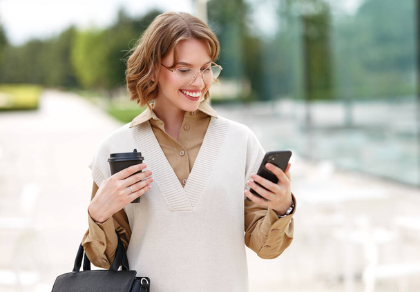 Портрет молодой очаровательной деловой женщины, идущей на улицу вдоль зеркального здания в солнечный день с чашкой кофе на вынос глядя с сладкой зубастой улыбкой на экран смартфона в руке - Фото, изображение