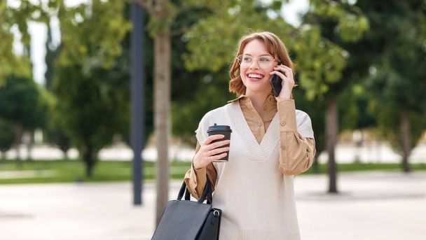 Young happy smiling female in glasses and formal outfit standing outside in city park with black handbag and cup of takeaway coffee while speaking on mobile phone with close friend or family - Zdjęcie, obraz