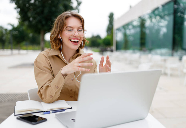 Szczęśliwy młody entuzjasta nauczyciel kobieta nauczyciel freelancer w słuchawki siedzi przed laptopem na tarasie na zewnątrz emocjonalnie gestykulacji z jasnym uśmiechem wyjaśnia uczniom podczas lekcji online - Zdjęcie, obraz
