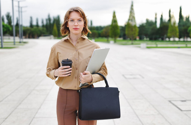 Confiant sérieux jeune femme d'affaires habillée en tenue de style d'affaires debout à l'extérieur avec ordinateur portable, tasse de café et sac à main classique noir, travailleuse de bureau posant sur fond de paysage urbain - Photo, image